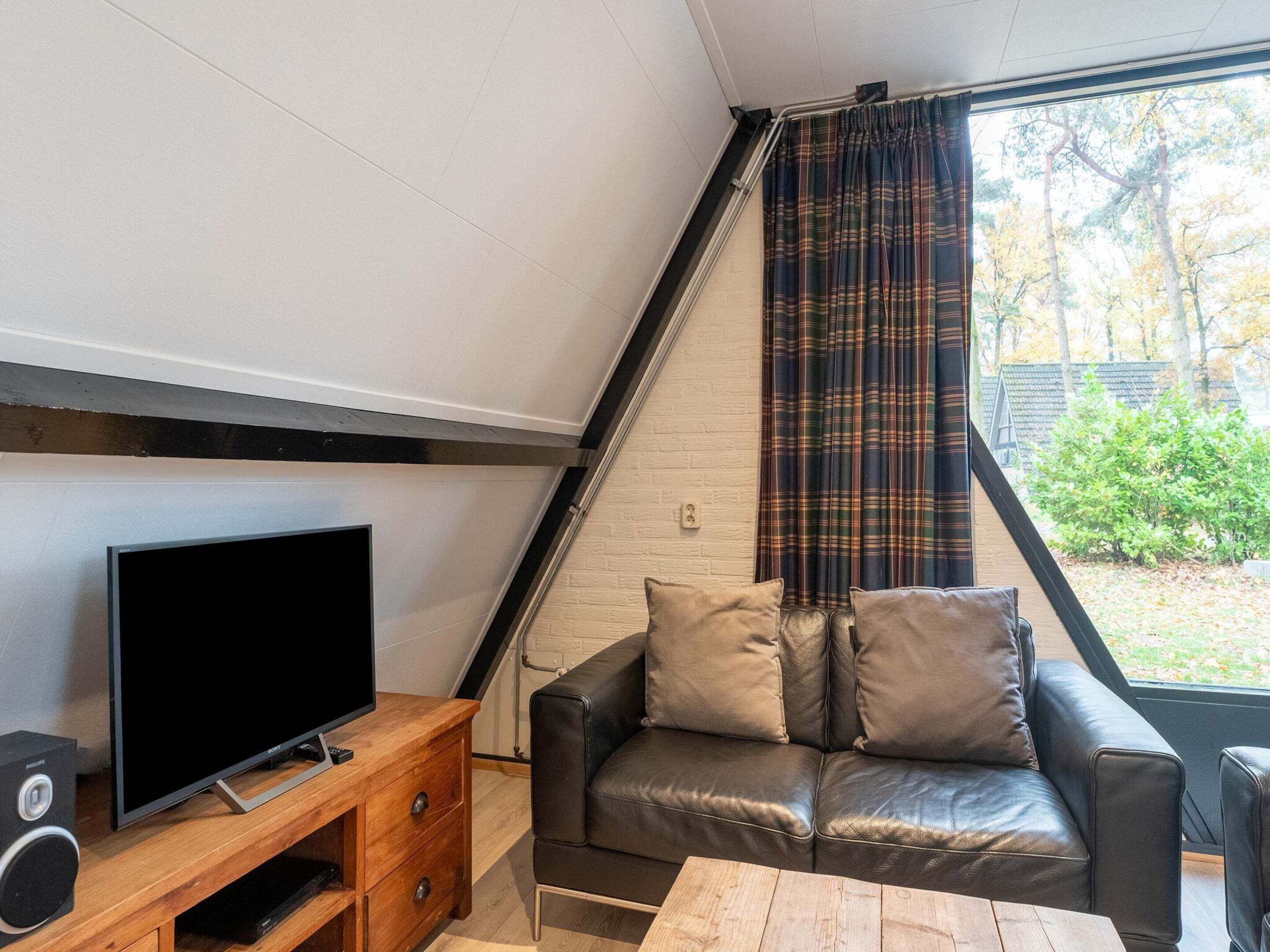 A cozy living room with a flat-screen TV on a wooden stand, a leather sofa with cushions, and a window with plaid curtains.