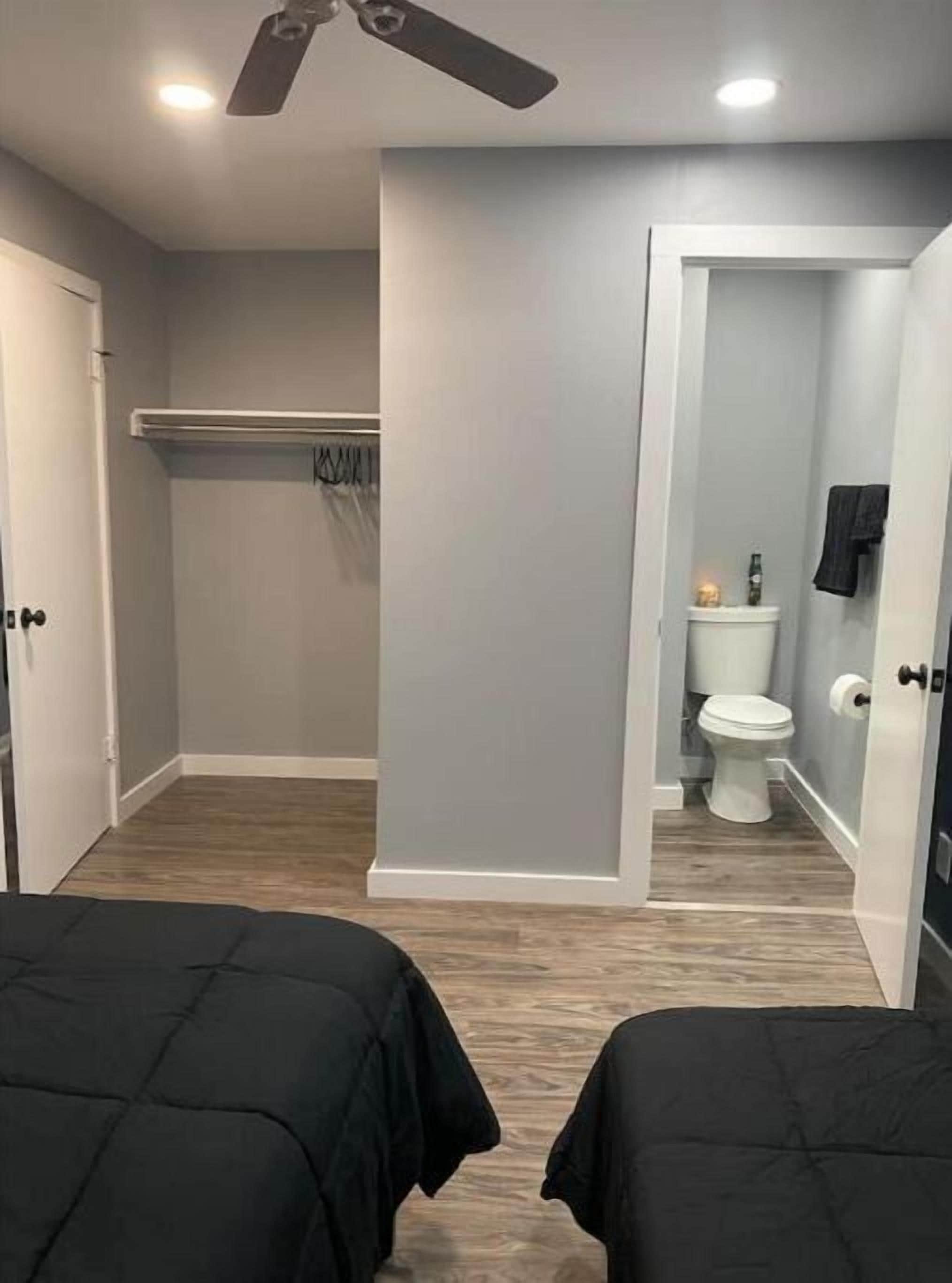 A room with two beds, a ceiling fan, a light fixture, a door, and a view into a bathroom with a toilet and towel rack.