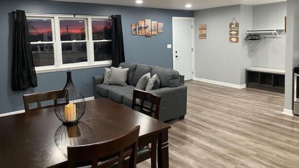 A modern living room with a grey sofa, a dining table with chairs, a large window with curtains, and a built-in bench with a coat rack. - Mankato’s Safe Haven (Mankato)