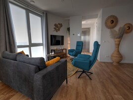 En moderne stue med en grå sofa, to blå lænestole, et træbord og et stort vindue med gardiner.