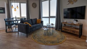 A modern living room with a sofa, coffee table, and TV. There is a dining area with chairs and a table, and a large window with curtains. - Apartment in Scherpenisse by Oosterschelde (Scherpenisse)