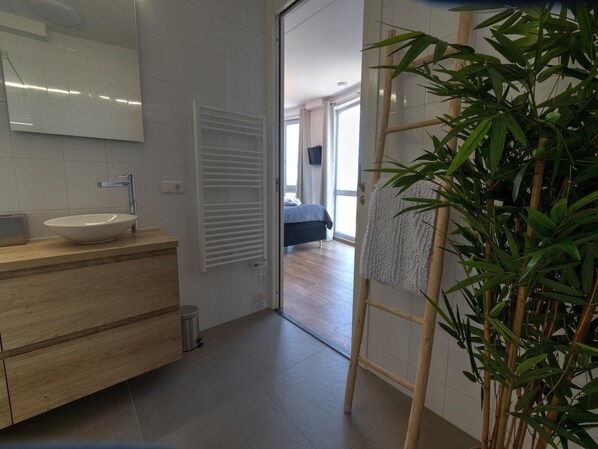 Une salle de bain moderne avec un lavabo blanc, un meuble-lavabo en bois et un radiateur. On peut voir un plant de bambou au premier plan.