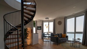 A modern living space with a spiral staircase, a dining area, a sofa with cushions, and large windows offering a view of the water. - Apartment in Scherpenisse by Oosterschelde (Scherpenisse)