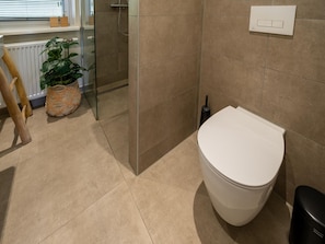 Une salle de bain comprenant des toilettes, un bidet, une douche en verre, une plante en pot et une chaise en bois.