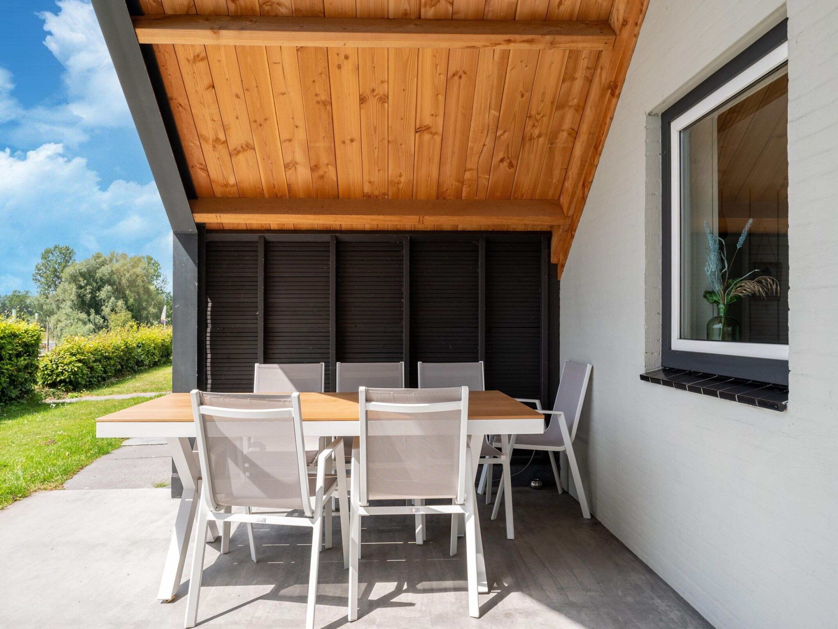An outdoor patio with a wooden table and white chairs, a window with a plant, and a view of greenery and trees.