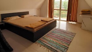 A bedroom with a bed, a window with curtains, a ceiling fan, and a small table with a lamp. - Gunjan Farms - Resort And Camping (Pune)