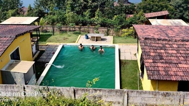 A swimming pool surrounded by buildings with red-tiled roofs, set against a backdrop of a lake and hills. - Gunjan Farms - Resort And Camping (Pune)