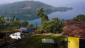 A view of a lake surrounded by hills, a parked car, and a yellow building with a tiled roof. - Gunjan Farms - Resort And Camping (Pune)