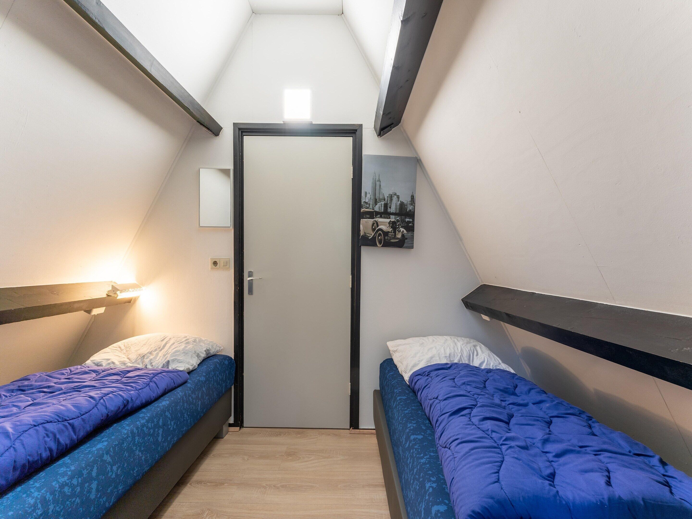 A small, narrow room with two single beds, a door, and a wall-mounted light fixture.