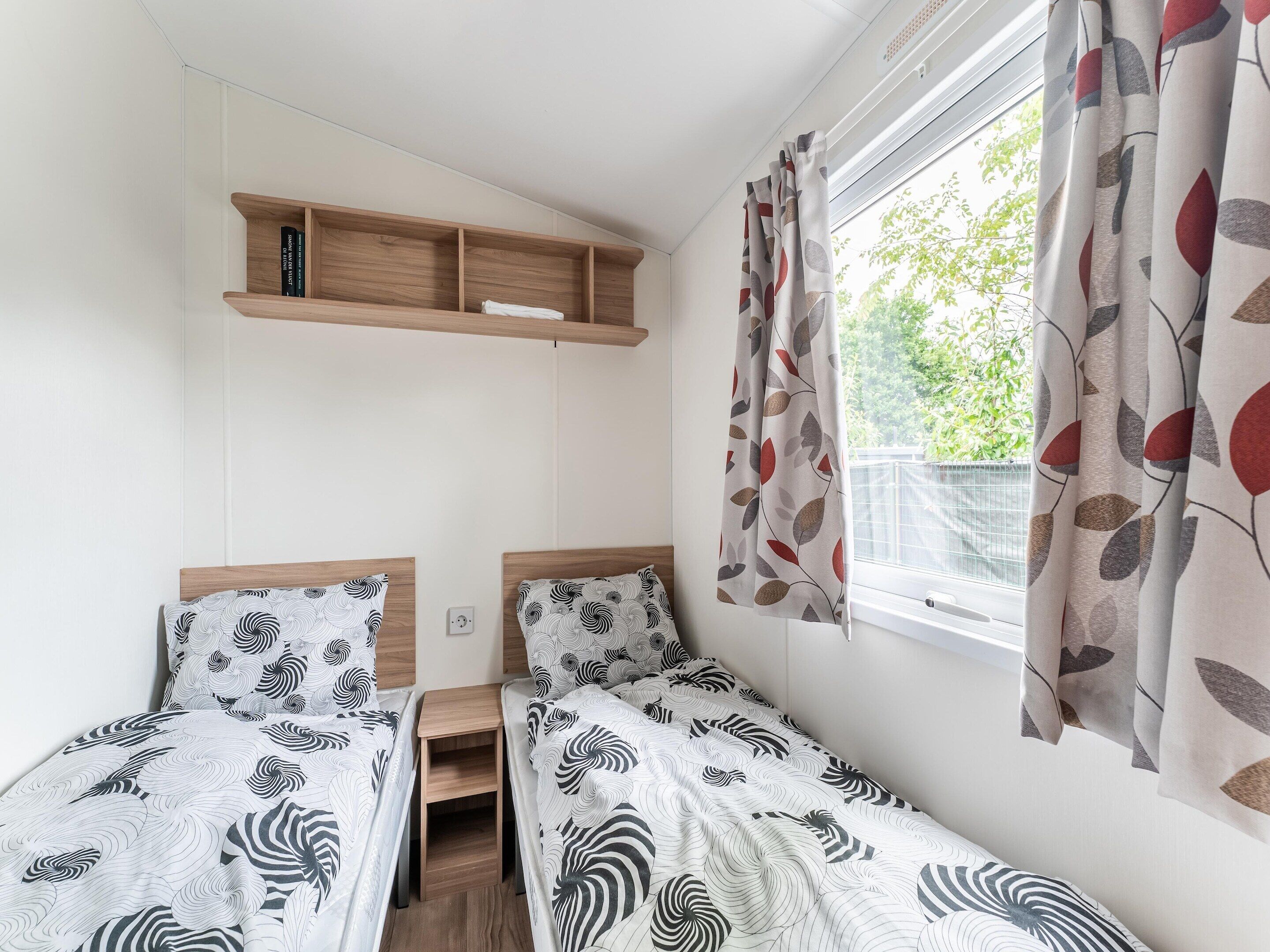 A bedroom with two single beds, a wooden shelf above, and a window with leaf-patterned curtains.