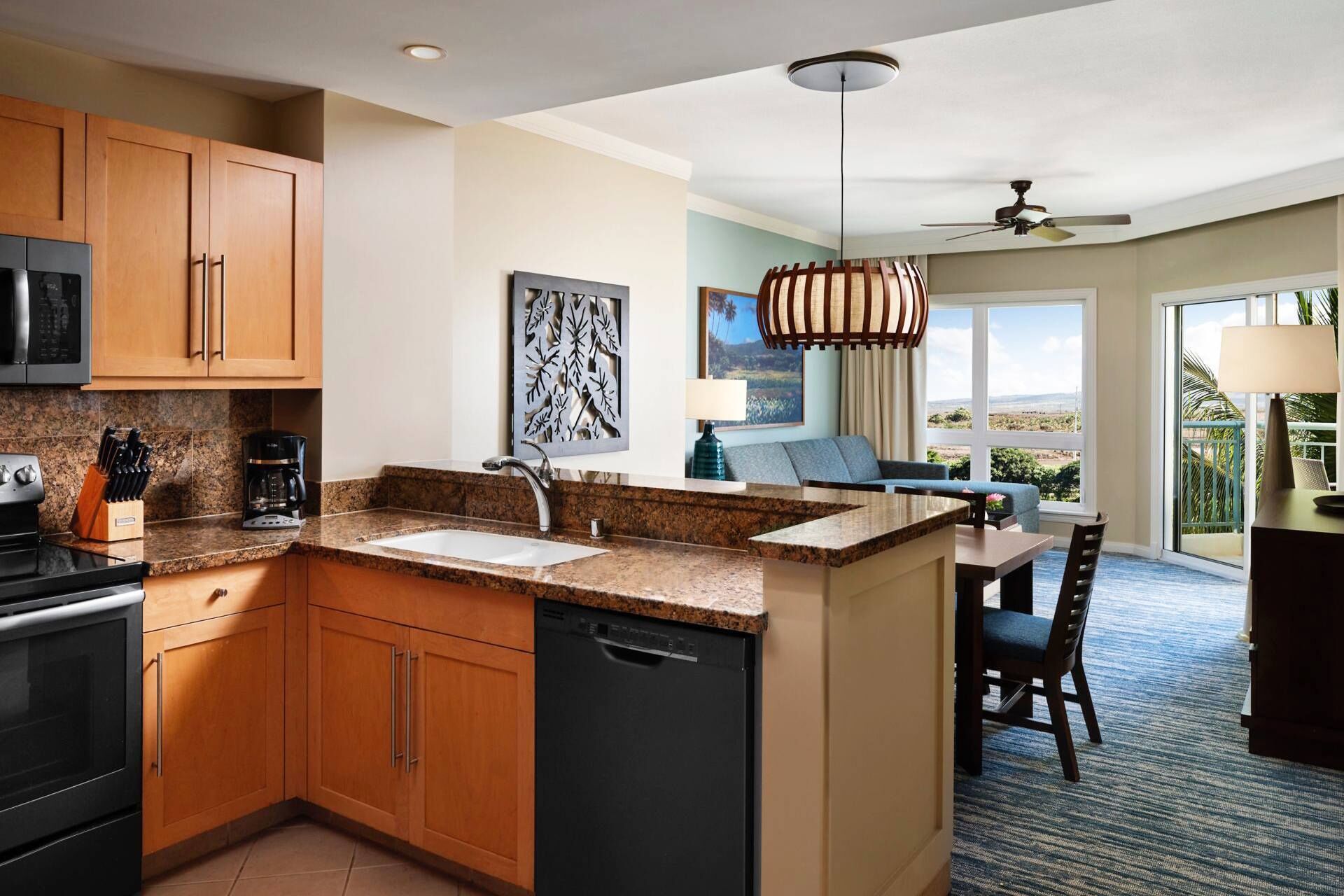 A modern kitchen with granite countertops, a dishwasher, and a built-in oven. There is a dining area with a table and chairs, and a view of the outdoors through large windows.
