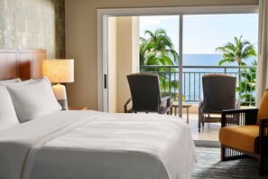 Een hotelkamer met een bed, een nachtkastje met een lamp, twee stoelen en uitzicht op de oceaan en palmbomen door een schuifdeur.