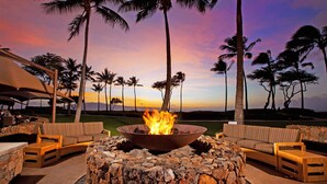 A stone fire pit surrounded by wicker furniture, set against a backdrop of palm trees and a sunset.