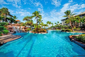 Relax by the clear pool surrounded by lush vegetation and palm trees, with a waterfall feature and scenic views from the resort's multi-story buildings.