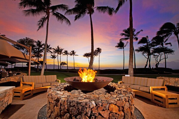 Relax in a tranquil outdoor space surrounded by palm trees and ocean views, featuring a central fire feature and comfortable seating.