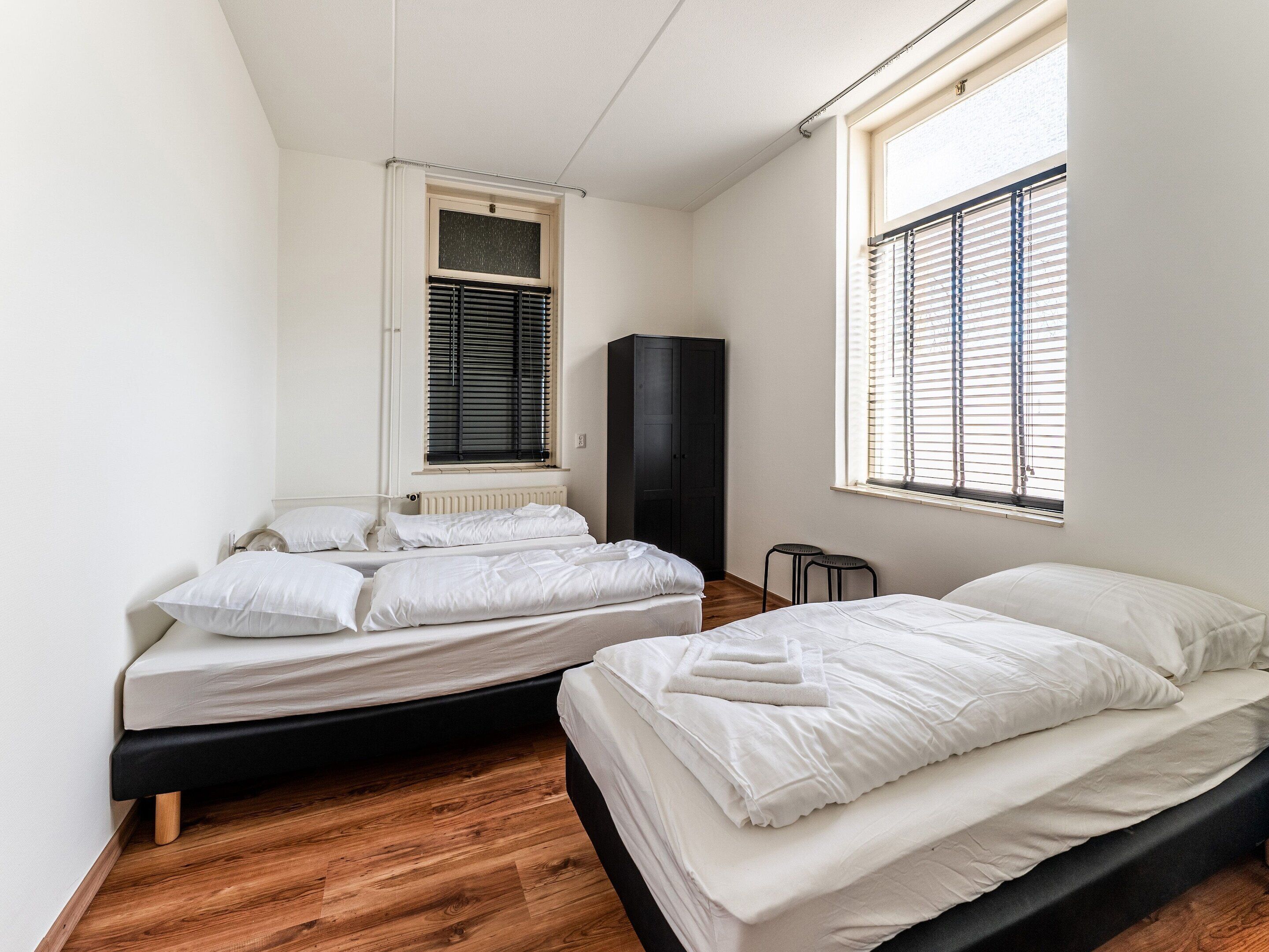 A bedroom with two beds, a radiator, a window with blinds, and a dark-colored wardrobe.
