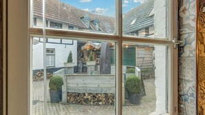 A window view of a courtyard with a wooden bench, potted plants, and a building with a tiled roof. - Beautiful Holiday Home in Voerendaal (Voerendaal)