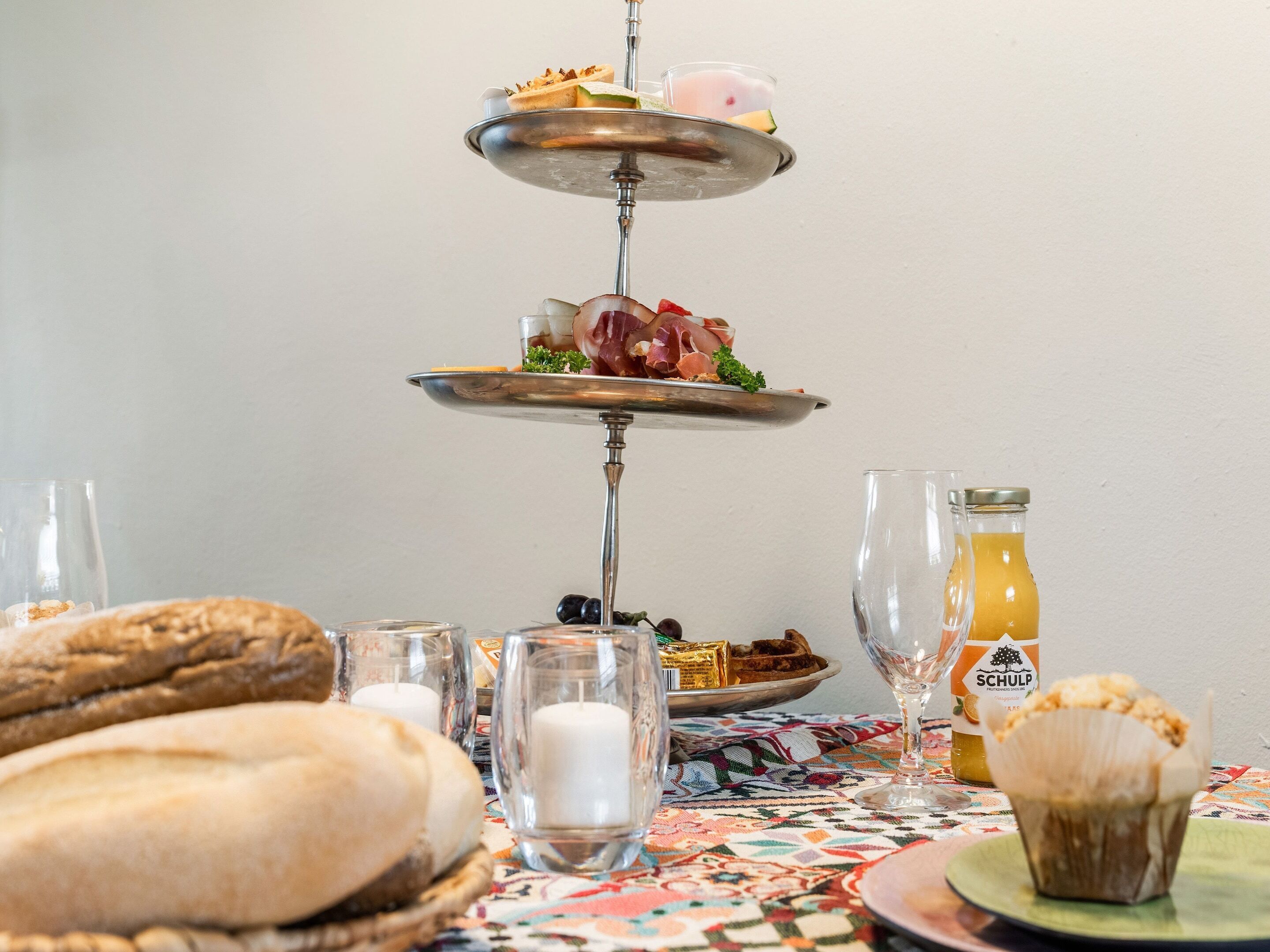 Una bandeja de tres niveles con comida y bebidas, un muffin, velas y una botella de jugo de naranja sobre una mesa con mantel estampado.