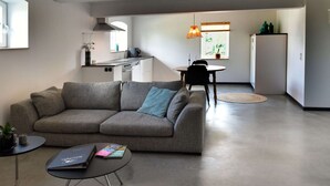 A modern living room with a grey sofa, a small coffee table, and a dining area with a table and chairs. A kitchen is visible in the background with a stove, sink, and cabinets. - Attractive Apartment in South Limburg (Klimmen)