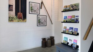 A room with white brick walls, a wooden door, and a shelf with travel brochures. There are two framed pictures on the wall, a shovel, and two metal containers on the floor. - Attractive Apartment in South Limburg (Klimmen)
