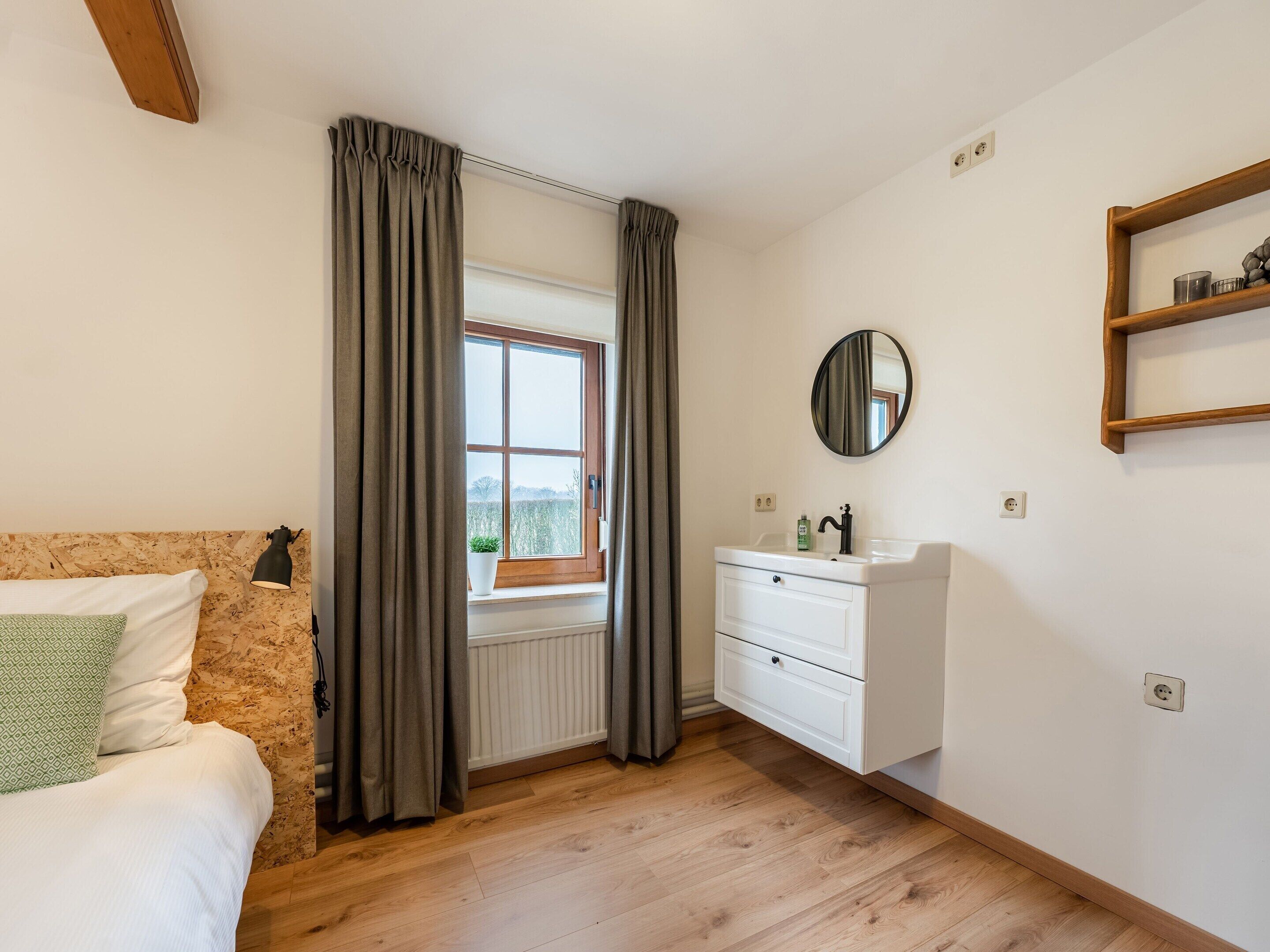 A bedroom with a wooden floor, a bed with a green pillow, a window with curtains, a bedside lamp, a white dresser with a mirror, and a shelf with a candle.