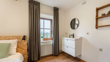 A bedroom with a wooden floor, a bed with a green pillow, a window with curtains, a bedside lamp, a white dresser with a mirror, and a shelf with a candle.