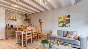 Un salon avec un canapé gris, des chaises en bois, une table basse et un coin cuisine équipé d’une cuisinière et d’étagères.