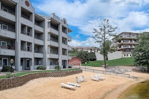 A multi-story residential building with balconies, a sandy beach area with lounge chairs, and a small brick structure.