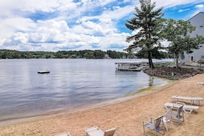 A lakeside beach with a dock, lounge chairs, and a large tree.