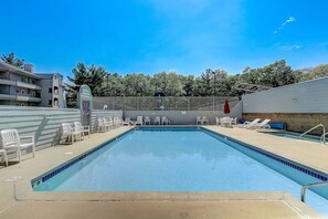 Une piscine rectangulaire à l’eau d’un bleu limpide, entourée de chaises longues blanches et d’un espace abrité sous un parasol rouge.