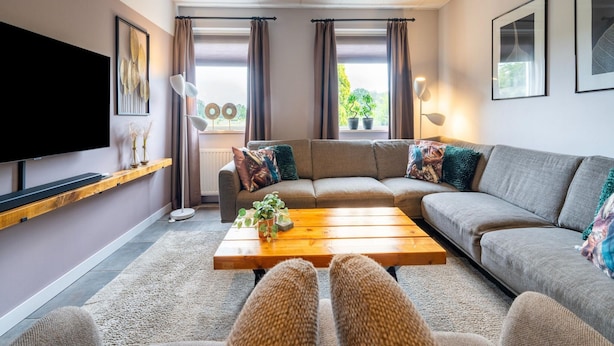 Un salon moderne avec un canapé d’angle, un téléviseur à écran plat, une table basse en bois et une grande fenêtre agrémentée de rideaux.