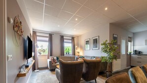 Un salon moderne avec un canapé, des fauteuils et un téléviseur. On y trouve également un coin cuisine équipé d’une machine à café et agrémenté d’une plante.