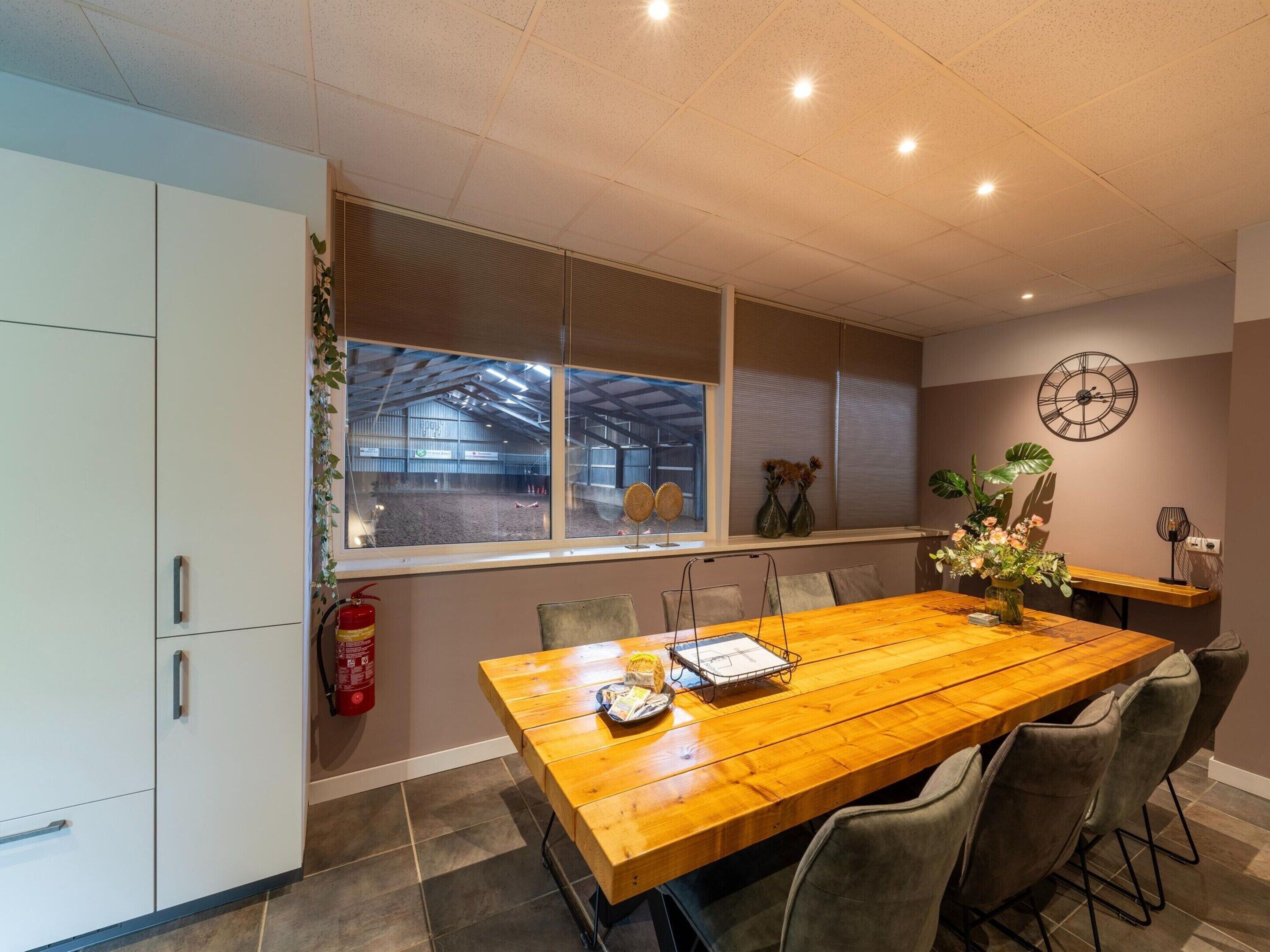 A dining area with a wooden table, chairs, a fire extinguisher, a window with blinds, and a wall clock.