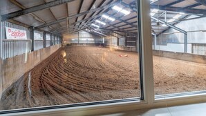 Une piste d’équitation intérieure avec un sol en terre, des poutres métalliques et un mur en verre.