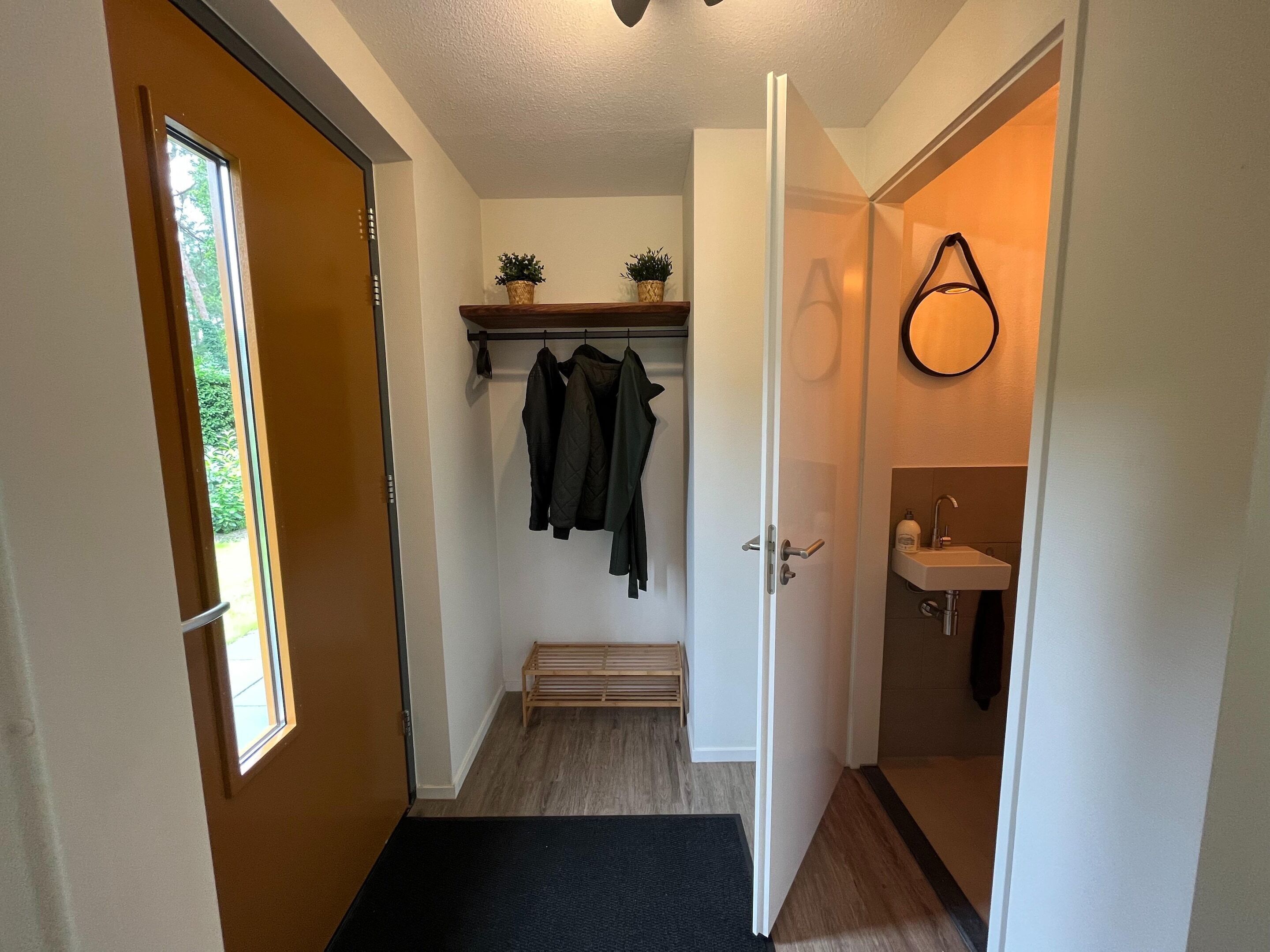A hallway with a wooden door, a bench, a coat rack with jackets, and a small shelf with plants.