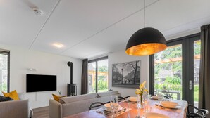 A modern living room with a fireplace, a dining area with a wooden table set for four, and a large window with a view of greenery. - Holiday Home in Lochem With Garden View (Lochem)