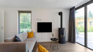 A modern living room with a sofa, a flat-screen TV, a black wood-burning stove, and a view of the outdoors through large windows. - Holiday Home in Lochem With Garden View (Lochem)