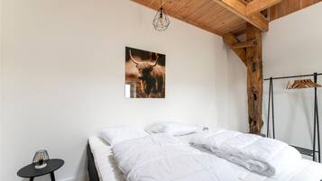 A bedroom with a wooden ceiling, a bed with white bedding, a small table with a candle, and a coat rack with hangers.