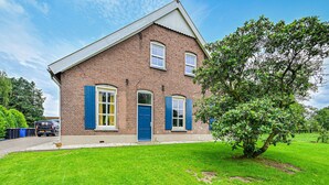Une maison à deux étages, construite en briques, avec une porte et des volets bleus, un petit jardin devant et un arbre.