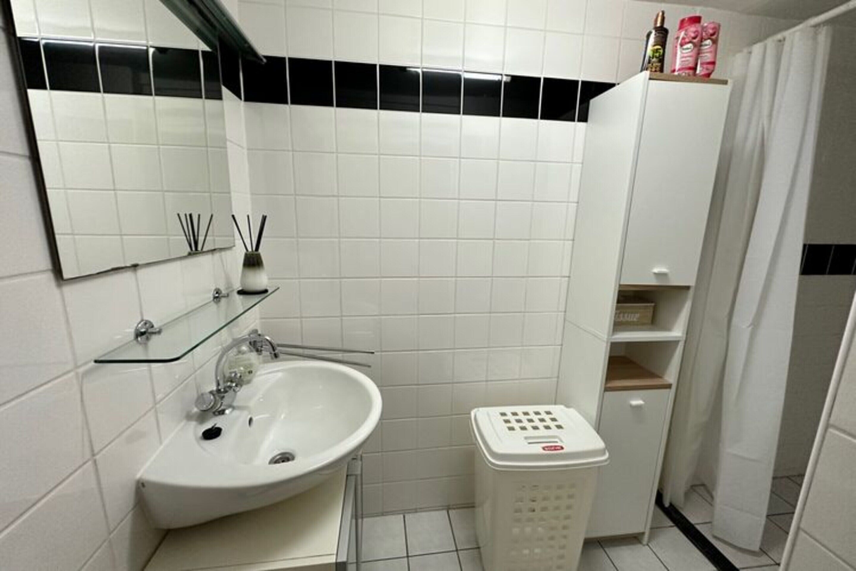 A bathroom with a white sink, mirror, and tiled walls. There is a glass shelf above the sink and a white laundry basket next to it.