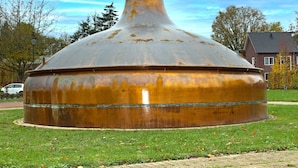 Une grande structure métallique rouillée, dotée d’un sommet arrondi et d’une base étroite, située sur une pelouse, avec un chemin pavé devant.