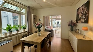 Un salon moderne avec une table à manger en bois, des chaises, un canapé et une table basse. Des plantes en pot sont posées sur le rebord de la fenêtre et une lampe sur la commode.