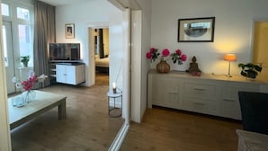 Un salon moderne avec un plancher en bois, un canapé blanc, une table basse et un téléviseur. On y trouve également un coin repas avec un banc et une table. Les murs de la pièce sont blancs et le plafond est doté d’un éclairage encastré.