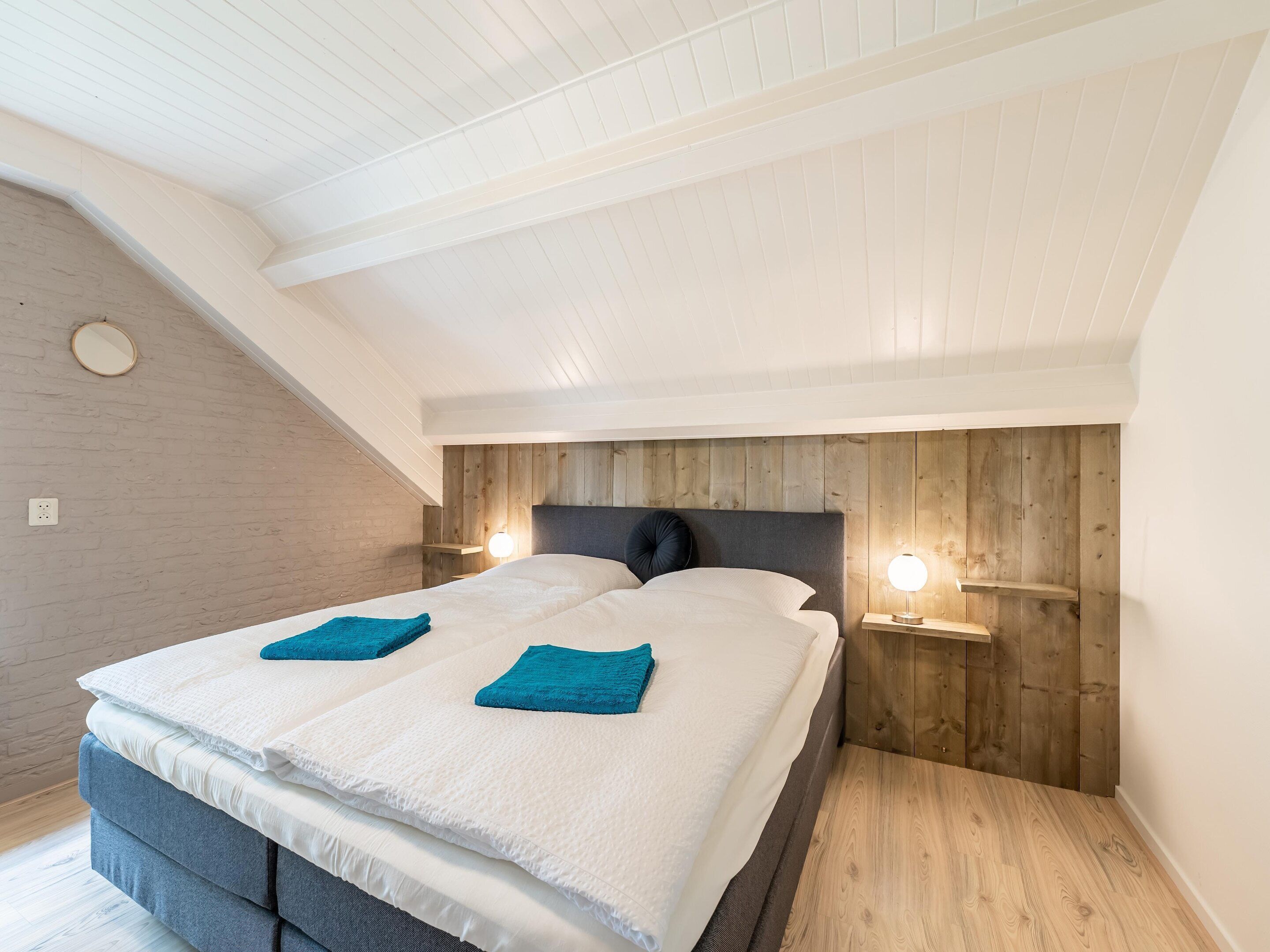 A bedroom with a sloped ceiling, wooden panel wall, two bedside tables with lamps, a bed with white bedding and teal pillows, and a grey sofa.