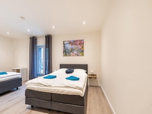 A modern bedroom with a large bed, a smaller bed, a nightstand, a lamp, and a window with curtains.