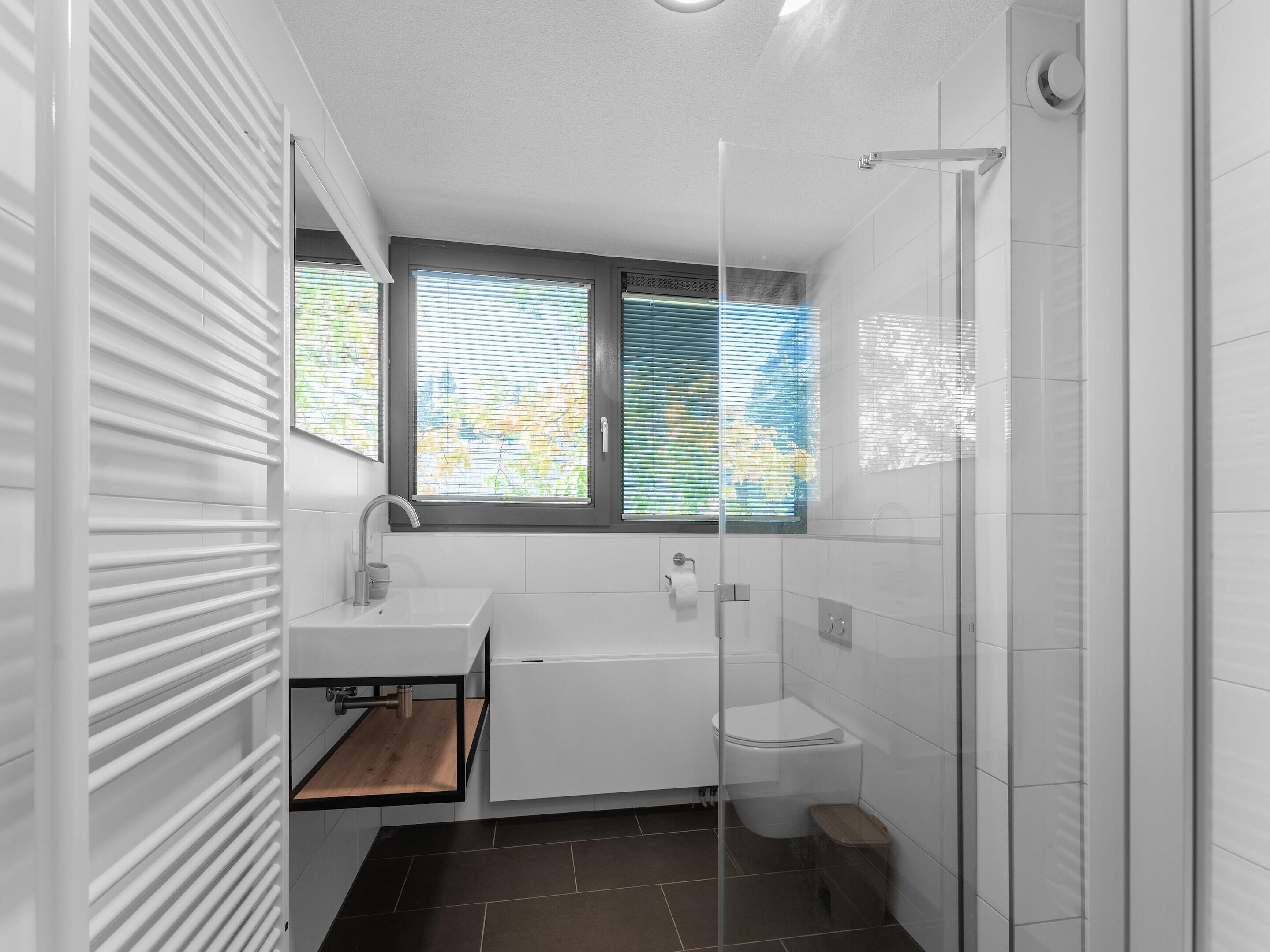 A modern bathroom with a white sink, a bathtub, and a shower. There are windows with blinds, and a wooden shelf under the sink.