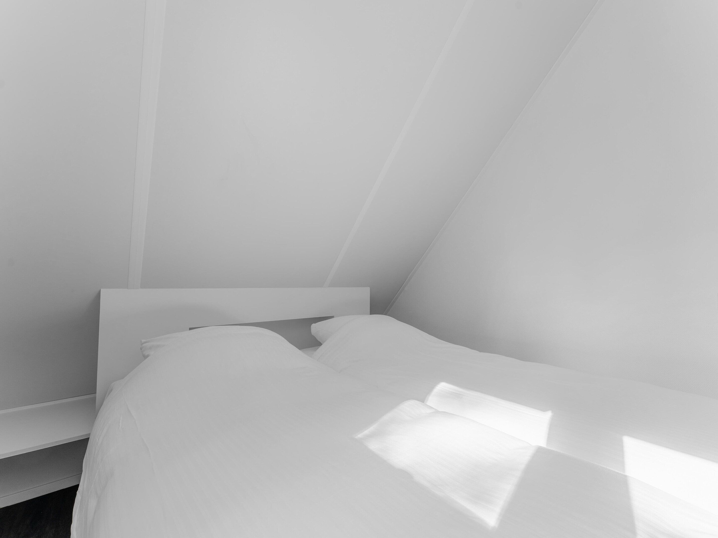 A minimalist bedroom with a low ceiling and a slanted roof, featuring a bed with white linens and a simple headboard.