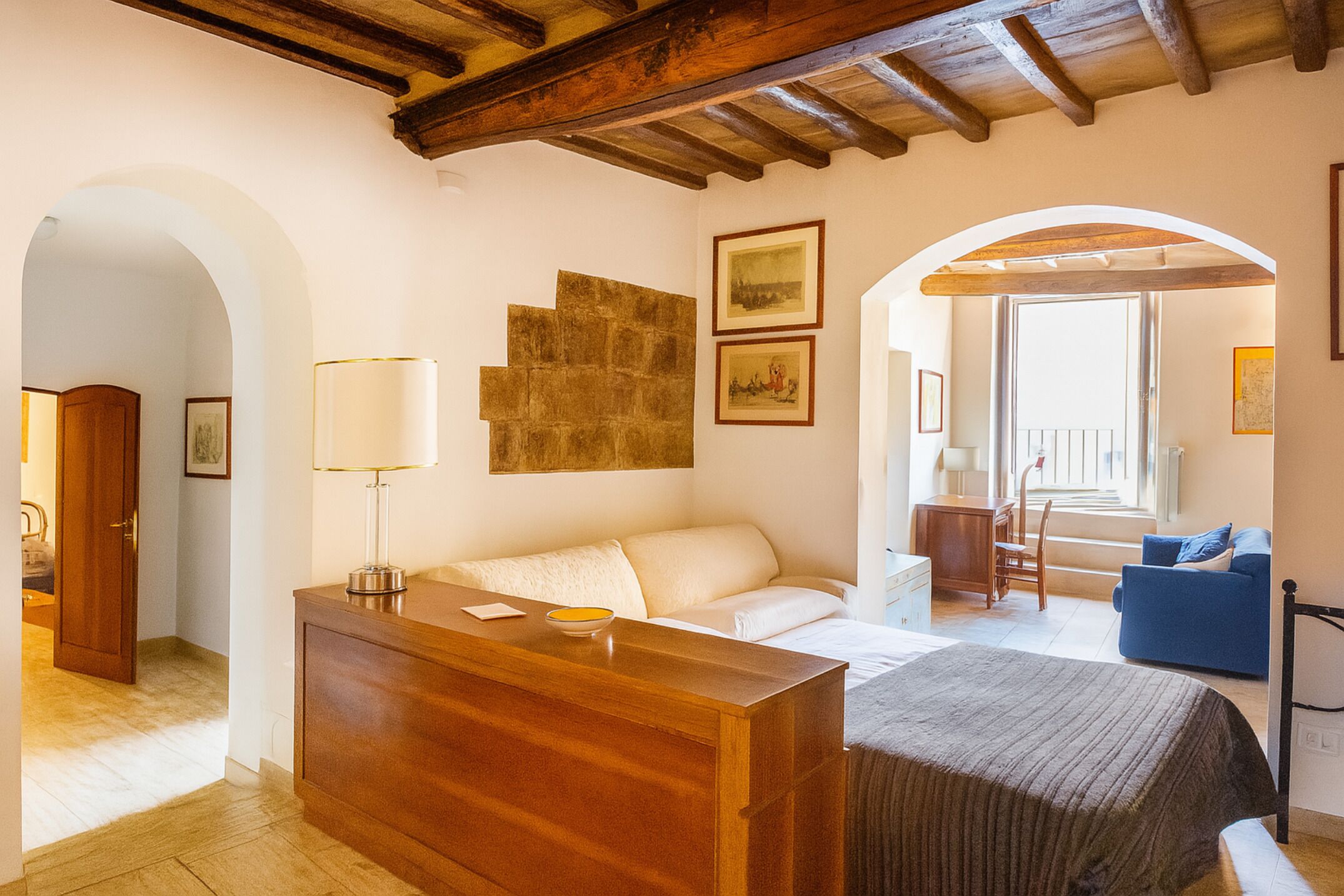 Ein Hotelzimmer mit einem Holztisch, einem weißen Sofa, einem blauen Sessel und einem Bett mit einer gestreiften Decke. An der Wand hängen zwei gerahmte Bilder und es gibt ein Fenster mit Blick.