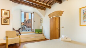 A bedroom with a bed, a chair, a desk with a computer, a window with a view of a building, and a door. - The Balcony Suite -Piazza del Campo View (Siena)