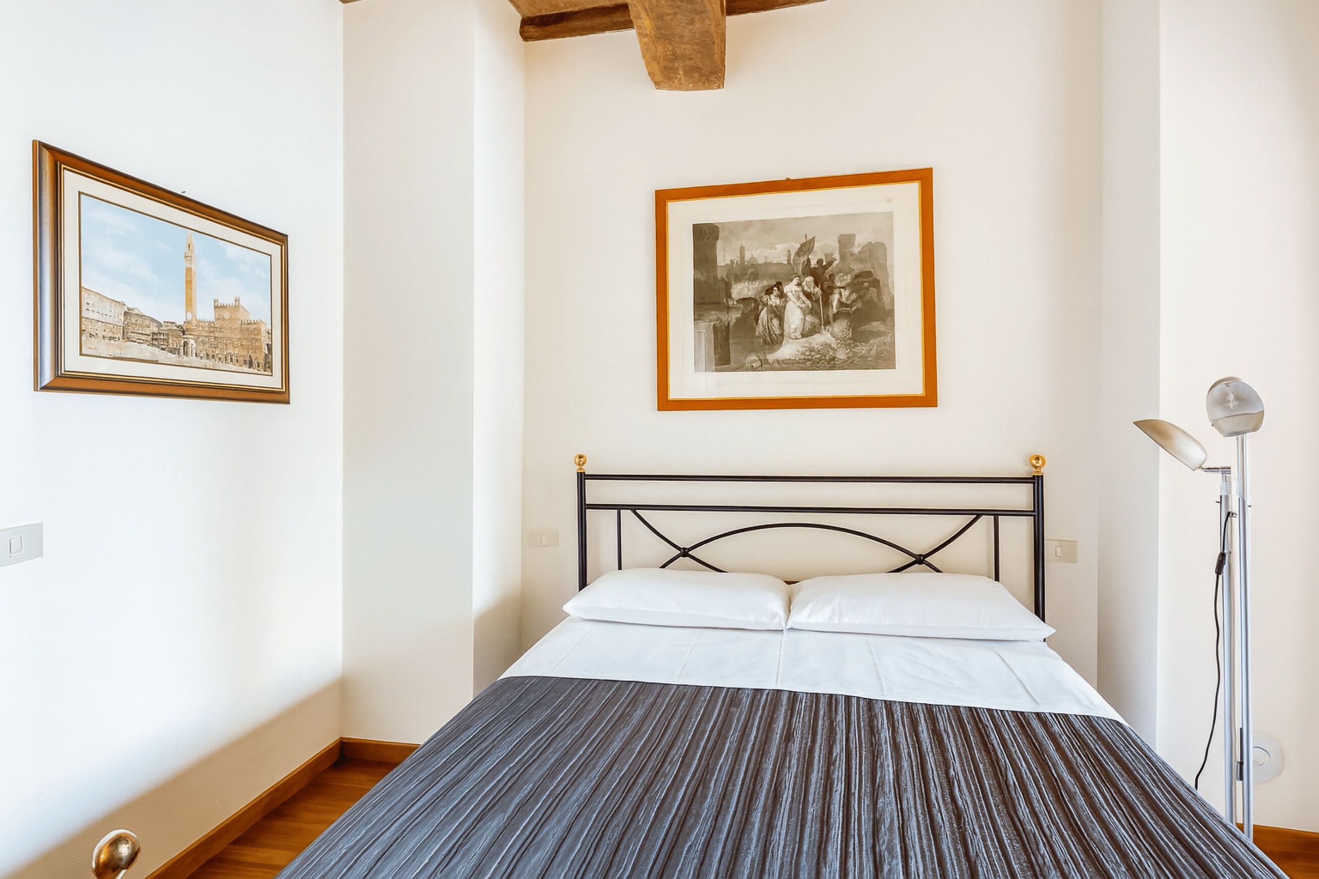A bedroom with a bed, framed pictures on the wall, and a floor lamp.
